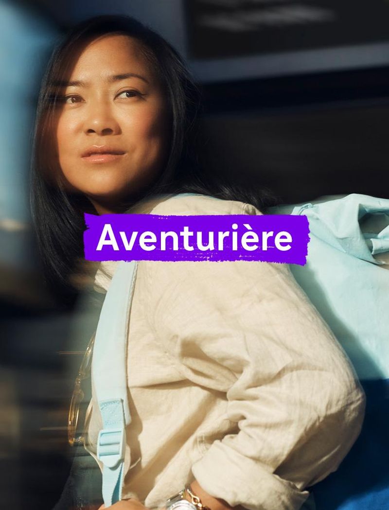 Une femme portant une chemise jaune et un sac à dos bleu regarde au loin avec une expression rêveuse. Le texte en surimpression indique : « Aventurière. »
