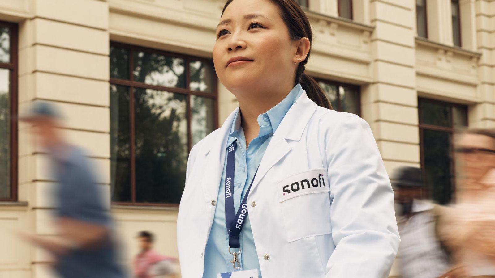 A woman in a white Sanofi lab coat and company badge outside of a building, moving forward while confidently and positively looking ahead. Several people are running alongside her in the background.