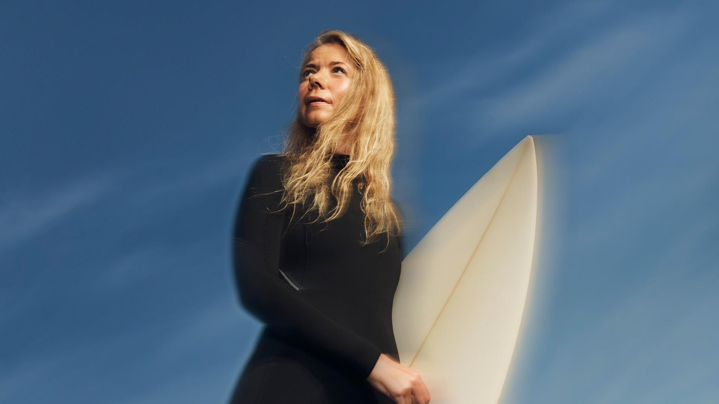 A woman wearing a black wetsuit holds a surfboard under blue skies and sunshine while looking out to the horizon with a hopeful and determined expression.