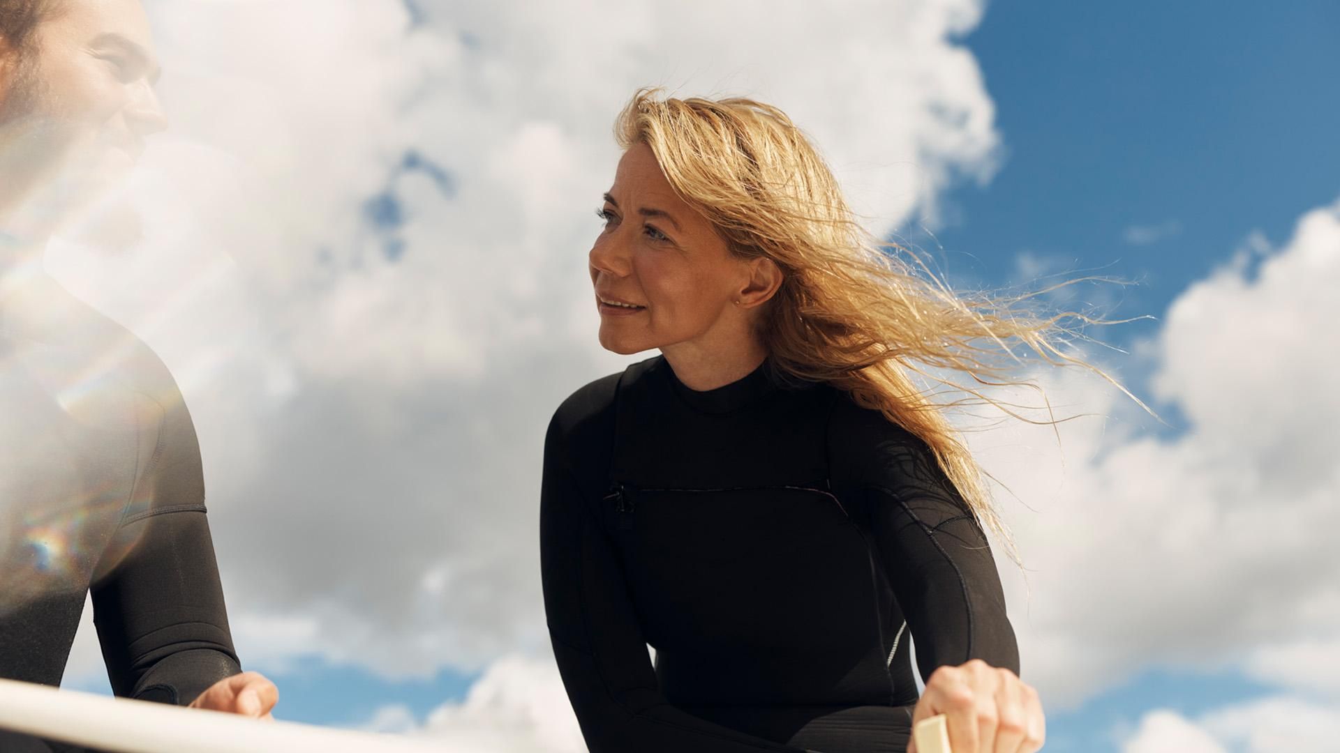A man and a woman wearing black wetsuits look at each other happily while applying wax to a surfboard in the sunshine, with clouds and blue skies in the background.​