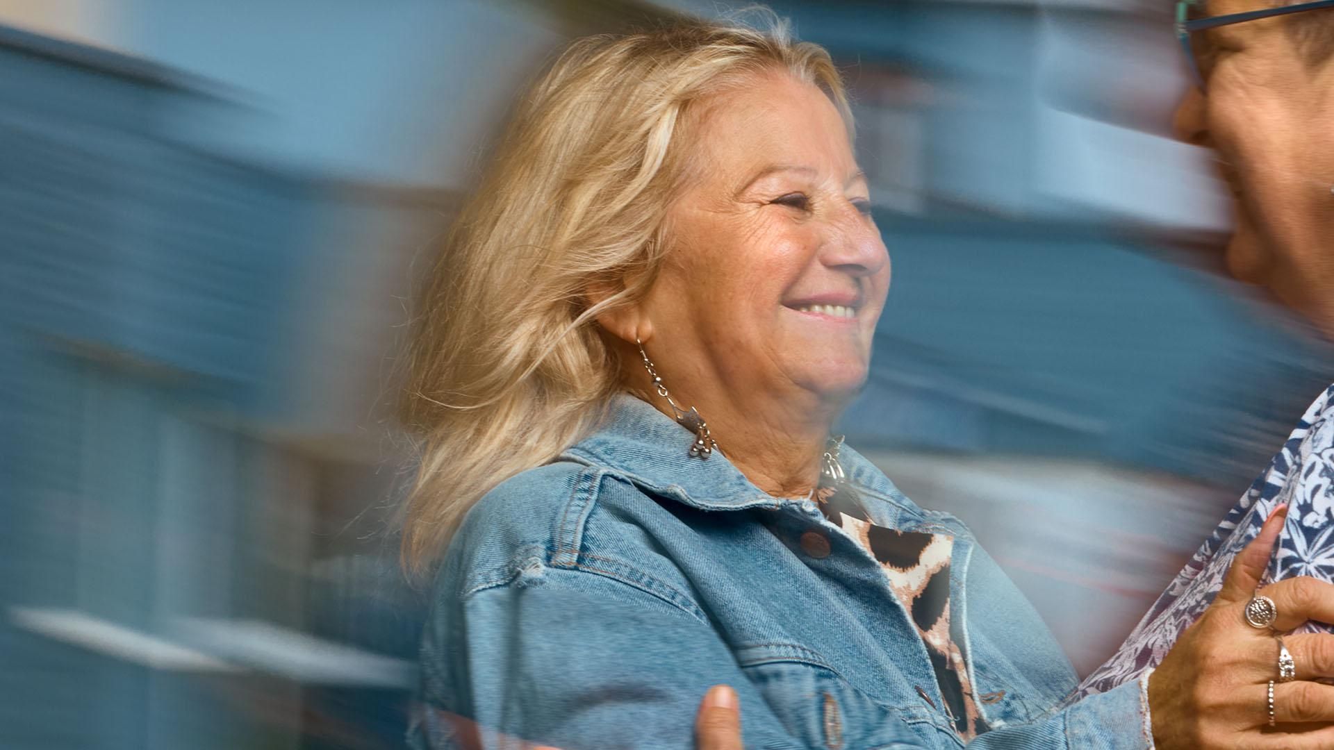 A smiling woman in a denim jacket reaches her hand out to embrace another woman who is smiling and wearing glasses. ​