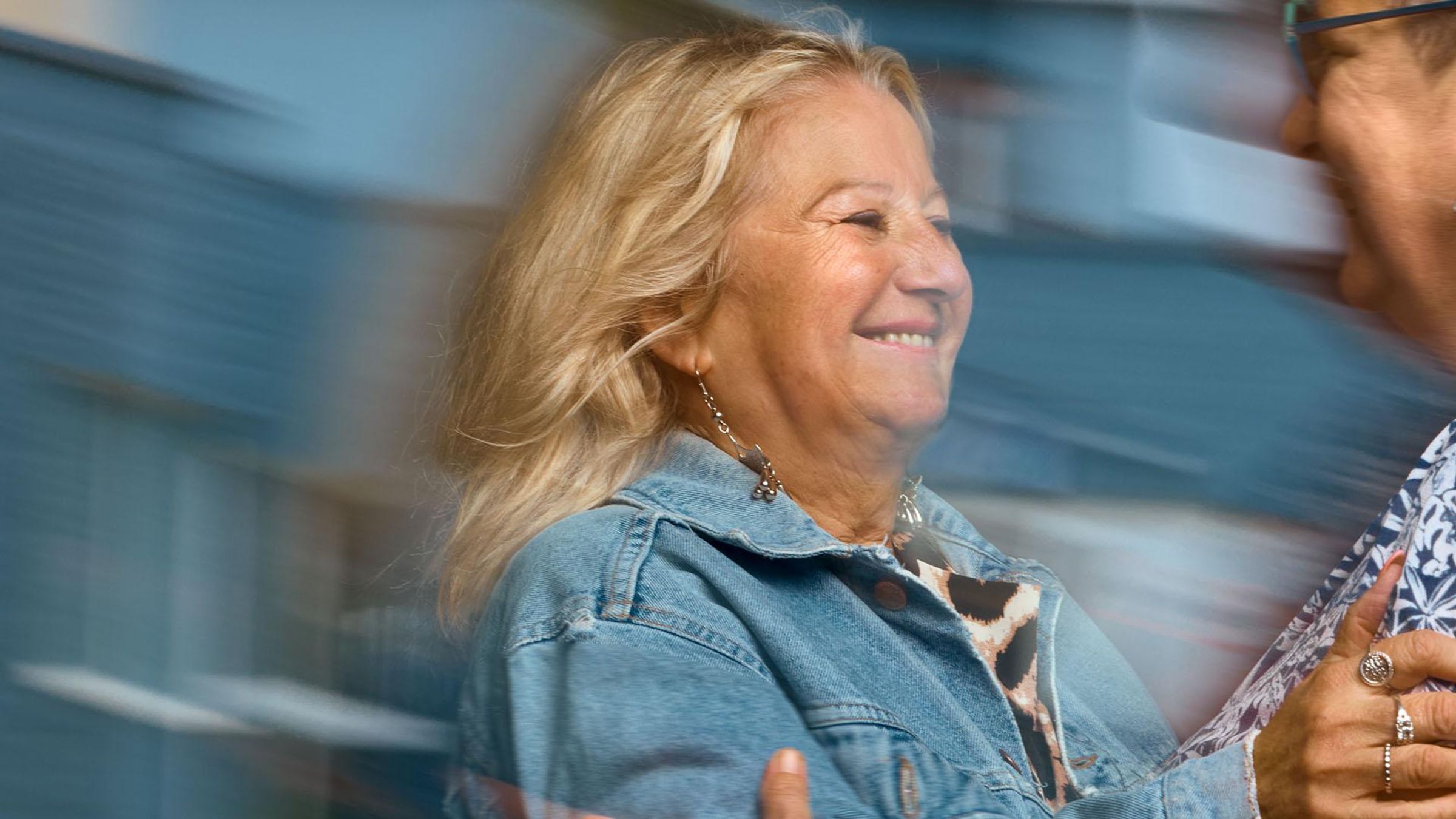 A smiling woman in a denim jacket reaches her hand out to embrace another woman who is smiling and wearing glasses. 