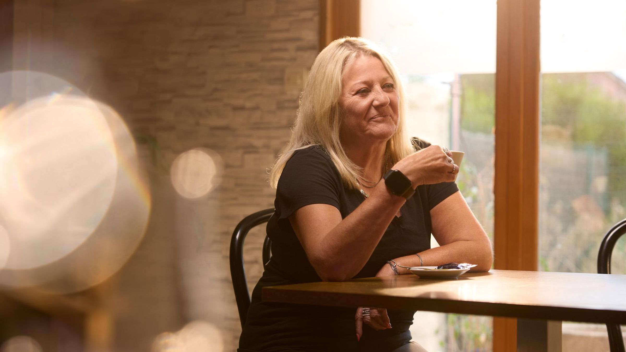 A smiling woman in a black t-shirt sits at a table in a sunlit café next to a window, holding a mug with a saucer beneath her.