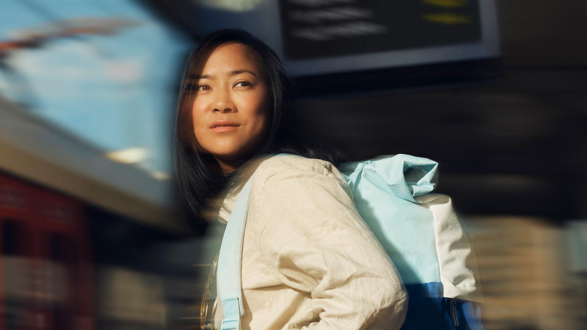A woman wearing a light yellow shirt and a blue backpack looks forward while walking through a train station. 
