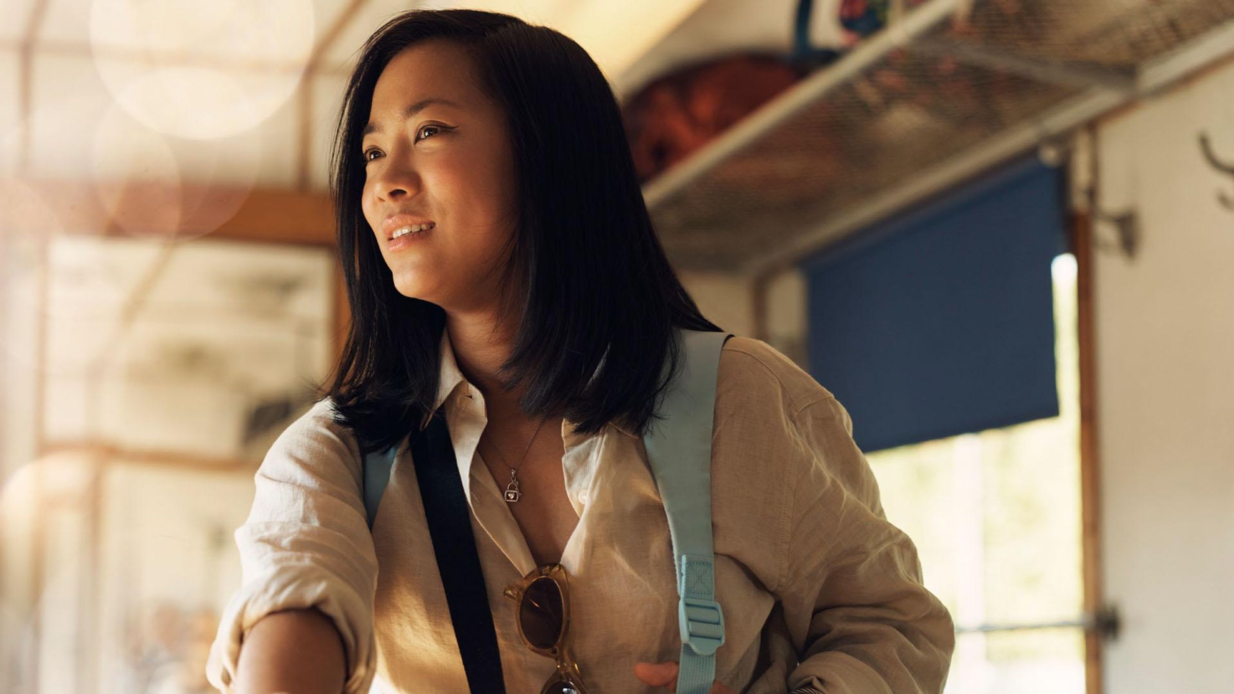 A woman wearing a light yellow shirt with a blue backpack and sunglasses hanging from her neck stands in the aisle of a bright train car, looking out of the window with a slight smile.