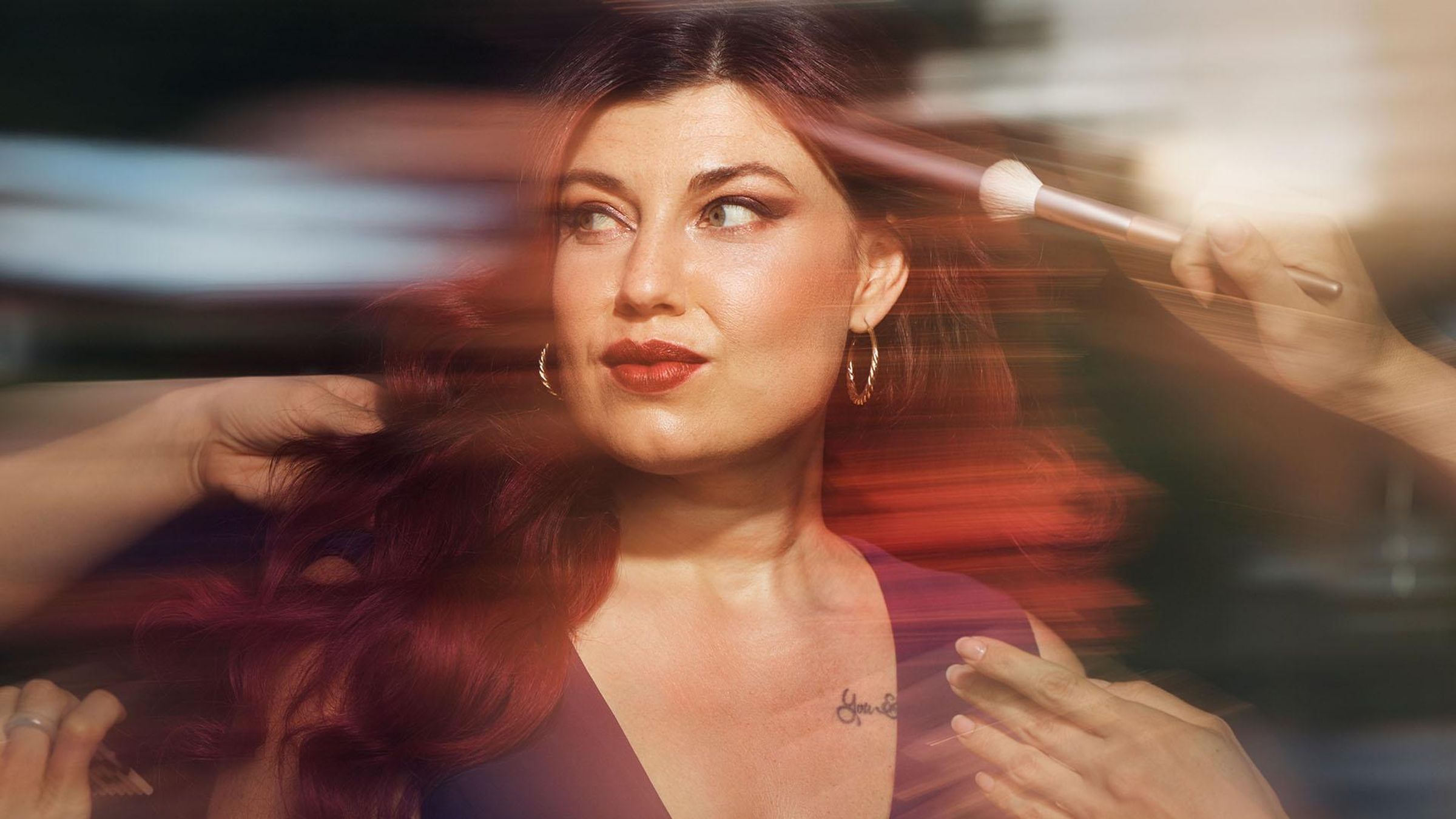 A glamorous woman wearing a purple dress is having her hair and makeup finished, she is looking to the side at someone as they fix her hair.