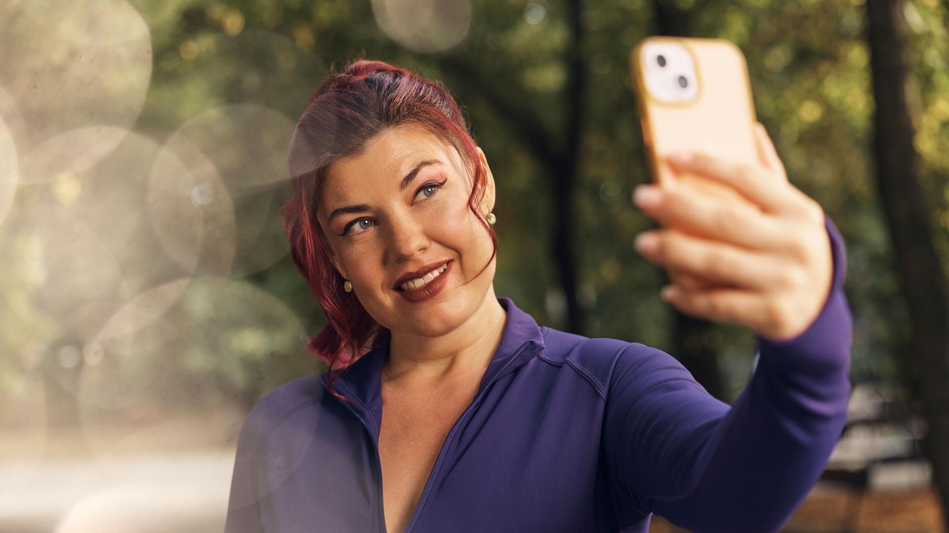 A smiling woman wearing a purple athletic top, holds up a smartphone to take a selfie outdoors. The background is blurred green foliage, and the sun is shining.​