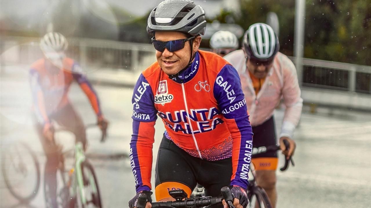 Group of cyclists racing on a wet road, with the lead cyclist wearing an orange and blue jersey with team branding.
