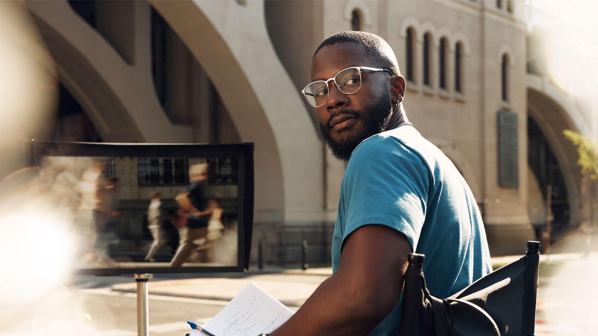 A man wearing glasses and a blue t-shirt sits in a director's chair and looks over his shoulder in the sunshine. He holds papers and a pen. In the background, a large monitor reflects blurred figures, and a bridge with arches is in the background.​