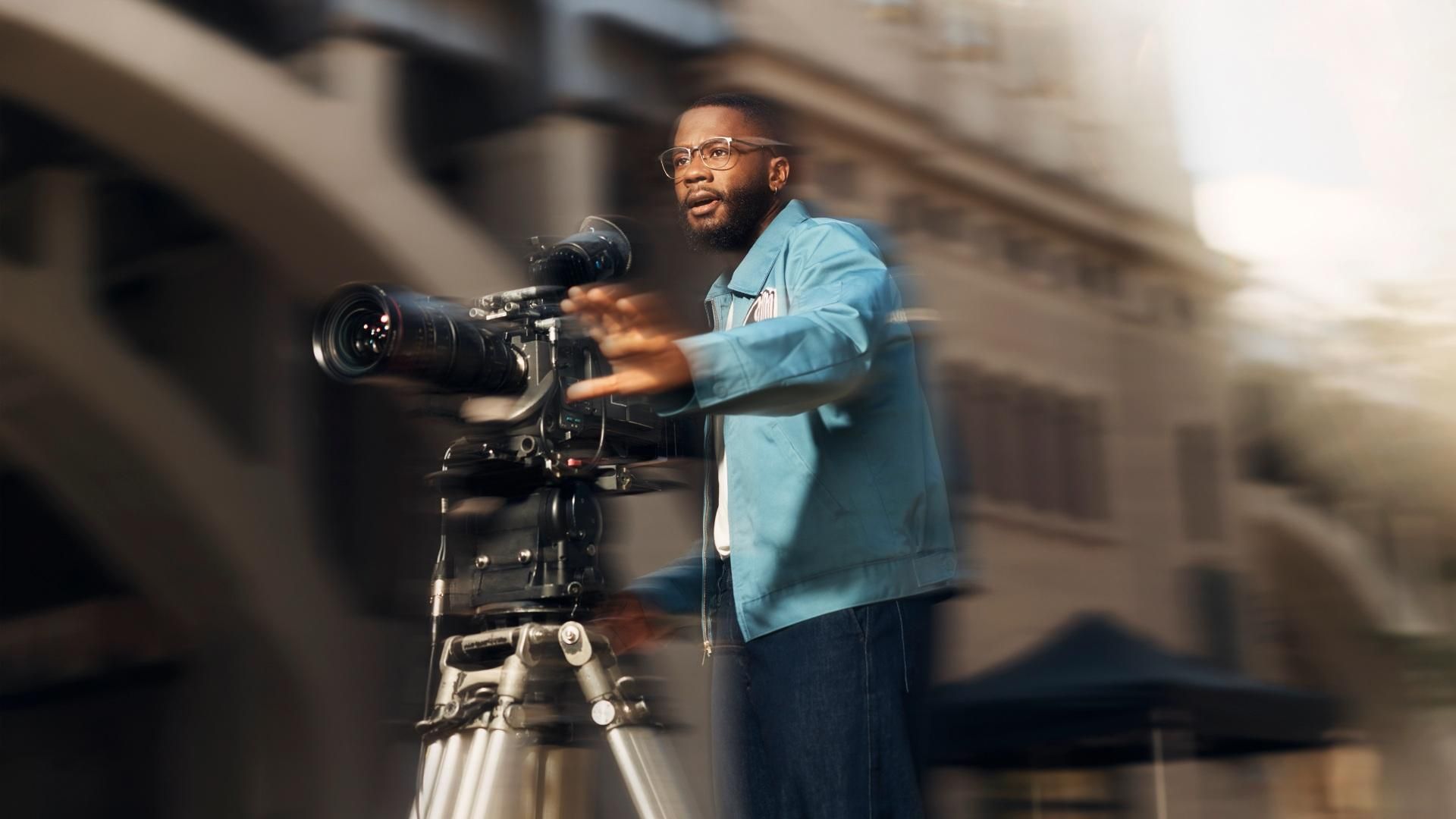 A man wearing glasses and a blue jacket operates a video camera and looks focused while filming, his hand is outstretched in front of him as he gestures. ​