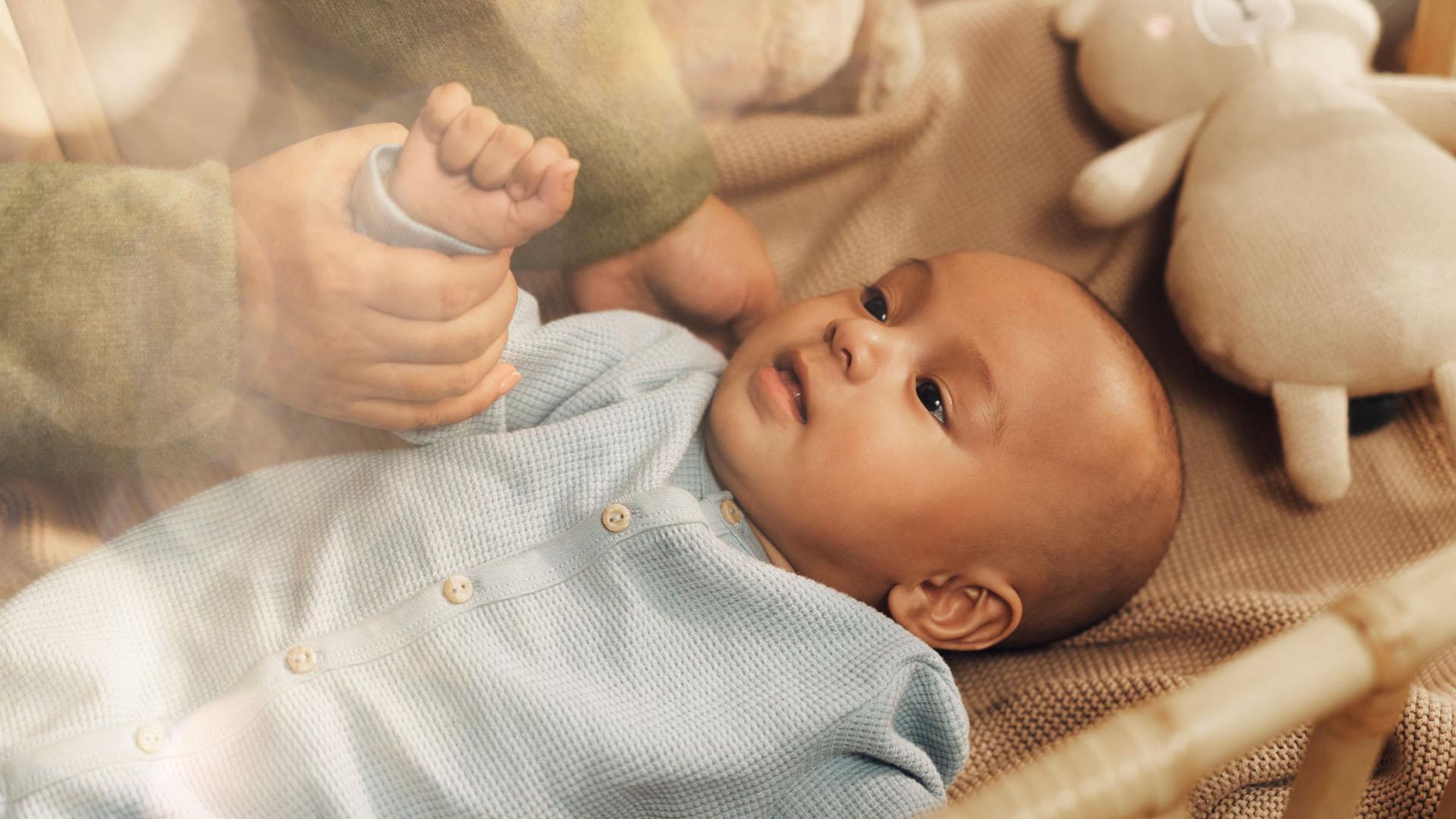 A small baby dressed in light blue clothing on a blanket in a crib is looking up. A woman’s hands are cradling the baby’s head and gently holding its arm, with a stuffed toy nearby.