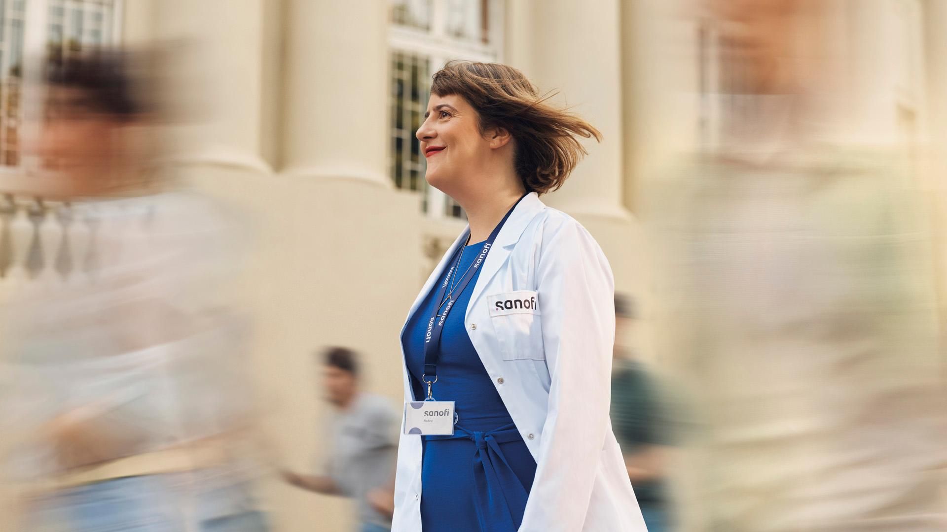 A smiling woman wearing a white Sanofi lab coat and company badge smiles while walking outdoors. Several people are running alongside her in the background, they are blurred in motion.​