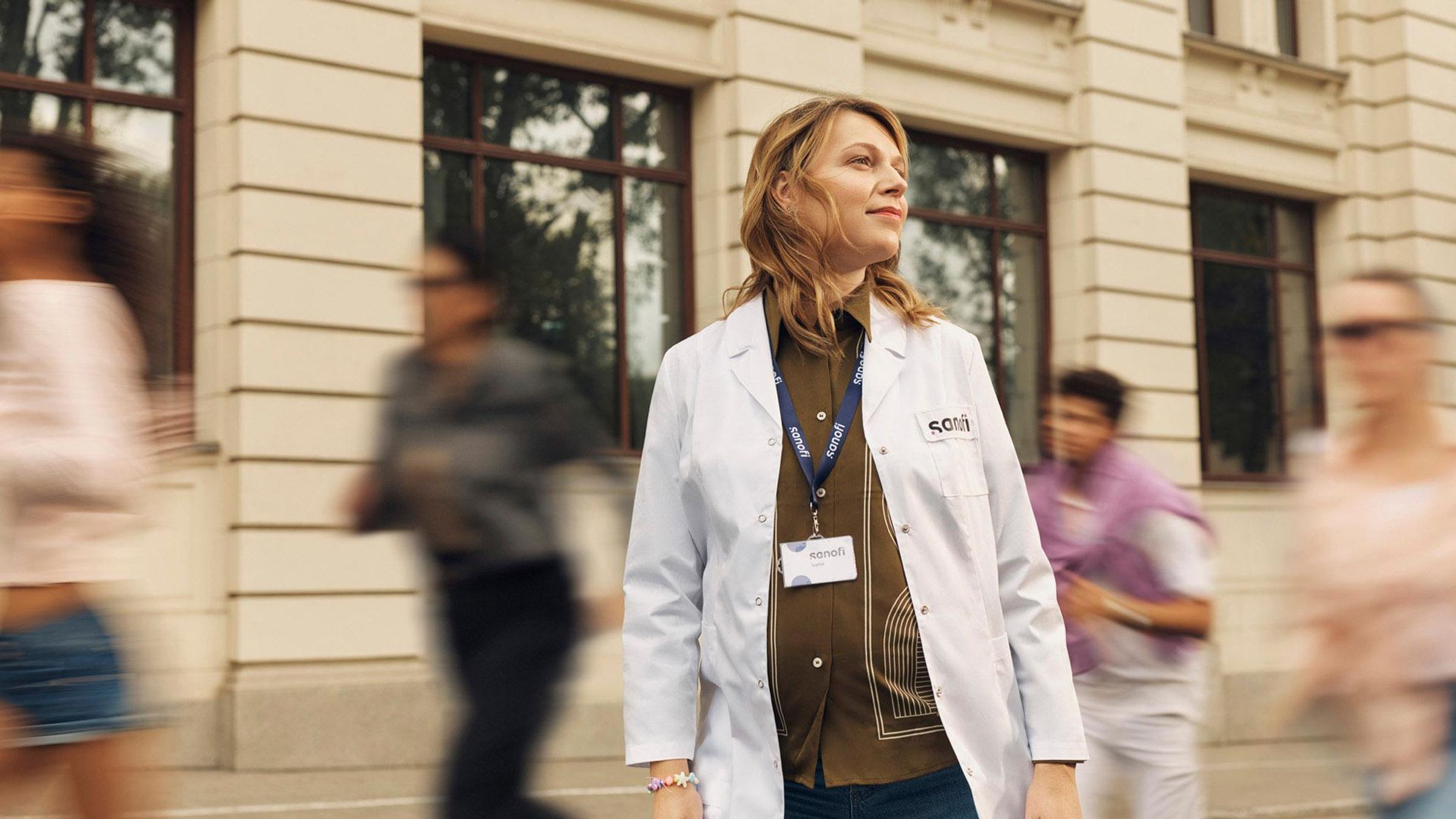 A woman in a white Sanofi lab coat and company badge looks ahead with an optimistic expression while standing outside a building. Several people are running alongside her in the background, they are blurred in motion.