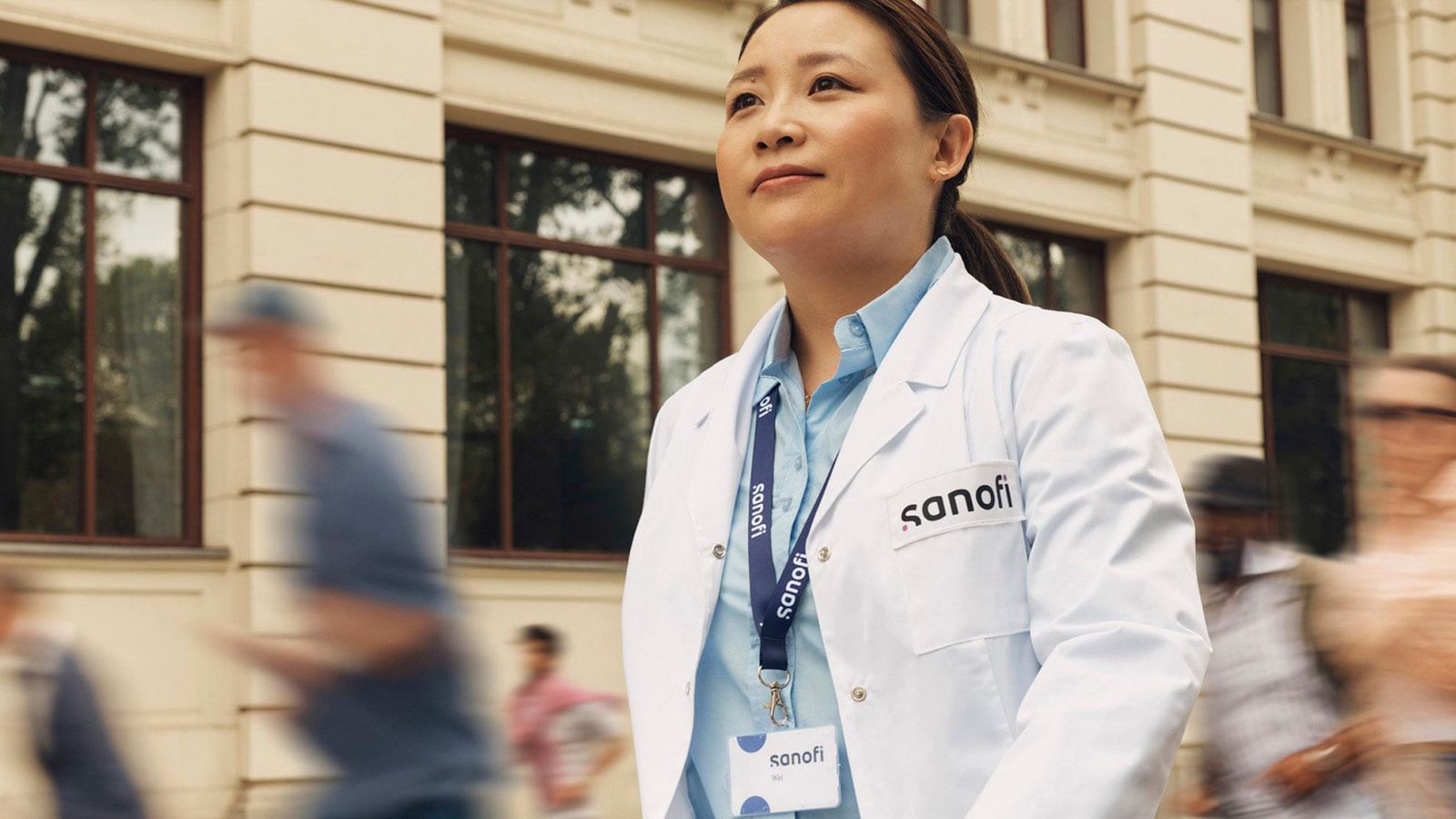 Image d'une collaboratrice Sanofi marchant dans la rue, elle porte un badge Sanofi et une blouse de scientifique