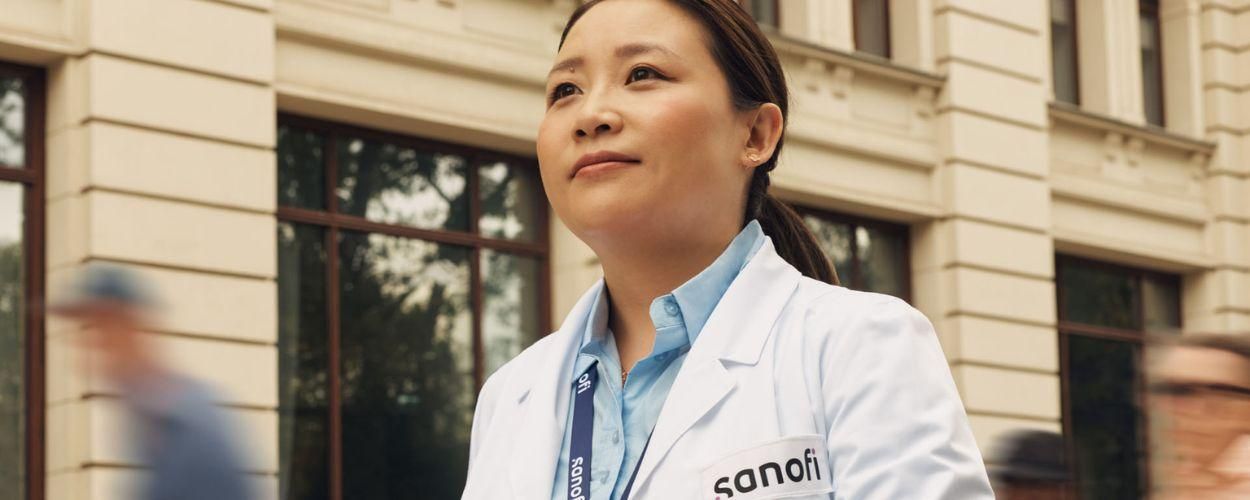 A woman in a white Sanofi lab coat and company badge outside of a building, moving forward while confidently and positively looking ahead. Several people are running alongside her in the background.​