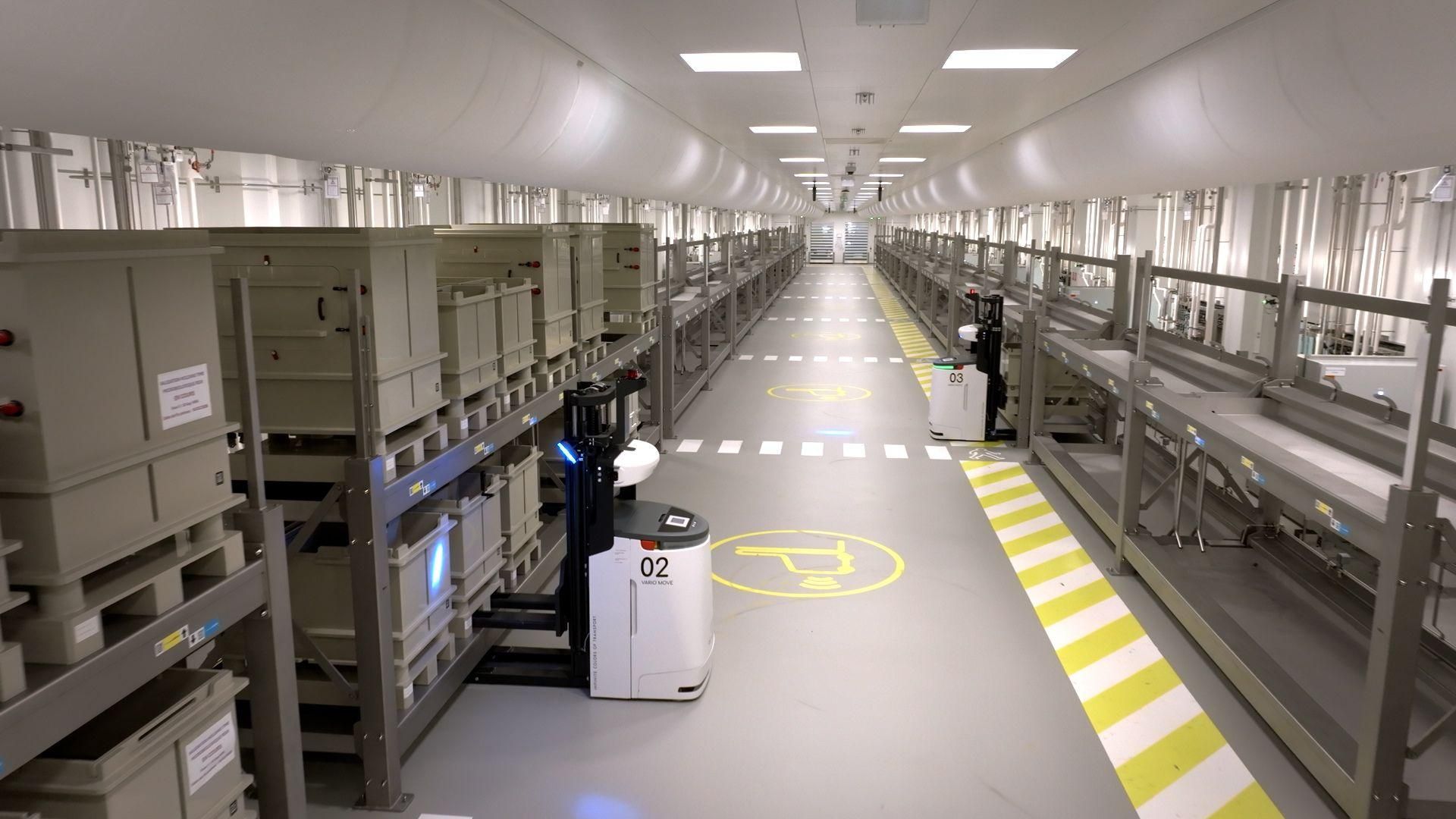 Two white and blue Automated Guided Vehicles (AGVs) navigating through controlled environment corridor at Sanofi Modulus in Neuville, France, lined with gray storage boxes on metal shelving, with yellow safety markings and robot-warning signs on the floor.