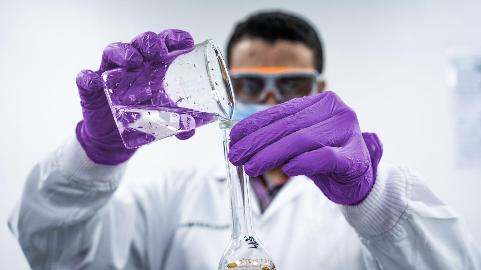 A man wearing purple gloves, white labcoat, and protective glasses, pours clear liquid from a beaker to a flask.
