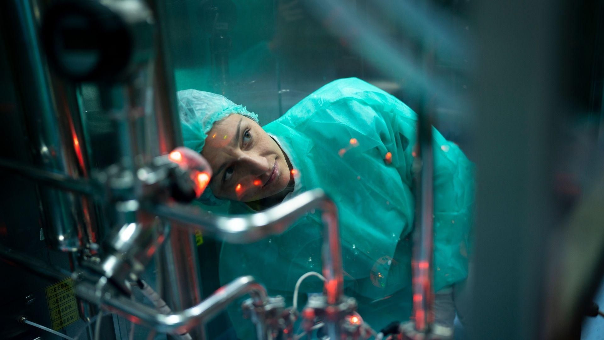 Loredana Ruscito, wearing a light green lab suit and hair net, looking up and through glass at lab equipment