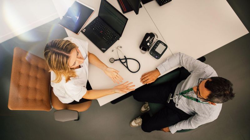 Overhead view of a desk with a healthcare professional and a patient in conversation, stethoscope and laptop visible.