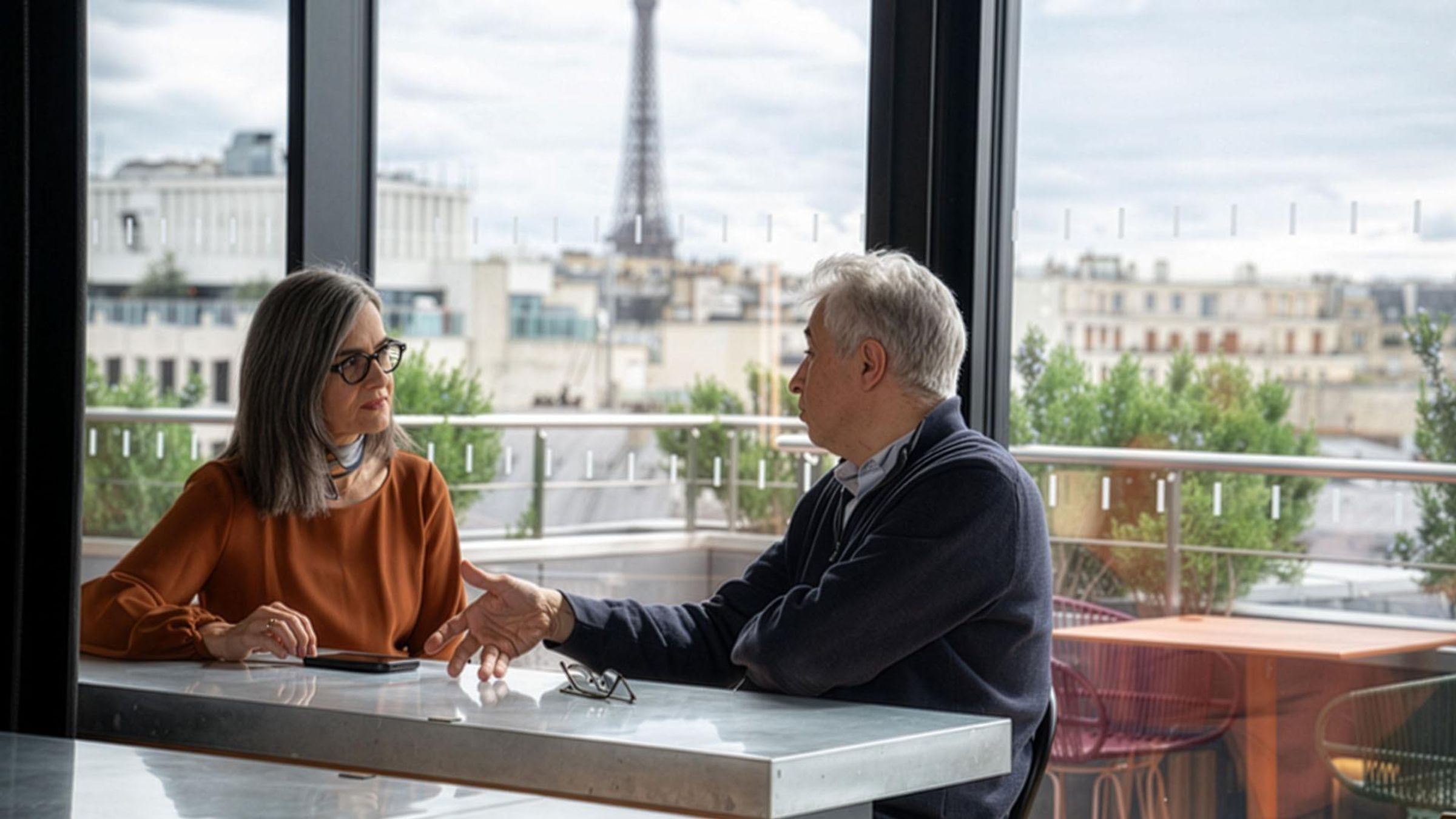 Two people sitting at a table indoors with a view of the Eiffel Tower through large windows in the background.