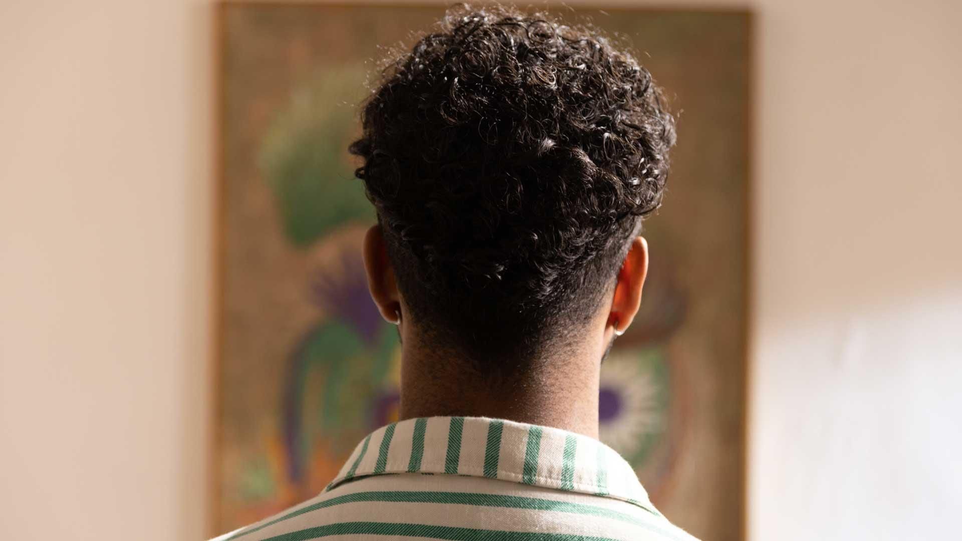The back of Bruno Araujo, standing with short curly hair and a white and green striped shirt, facing a painting on the wall