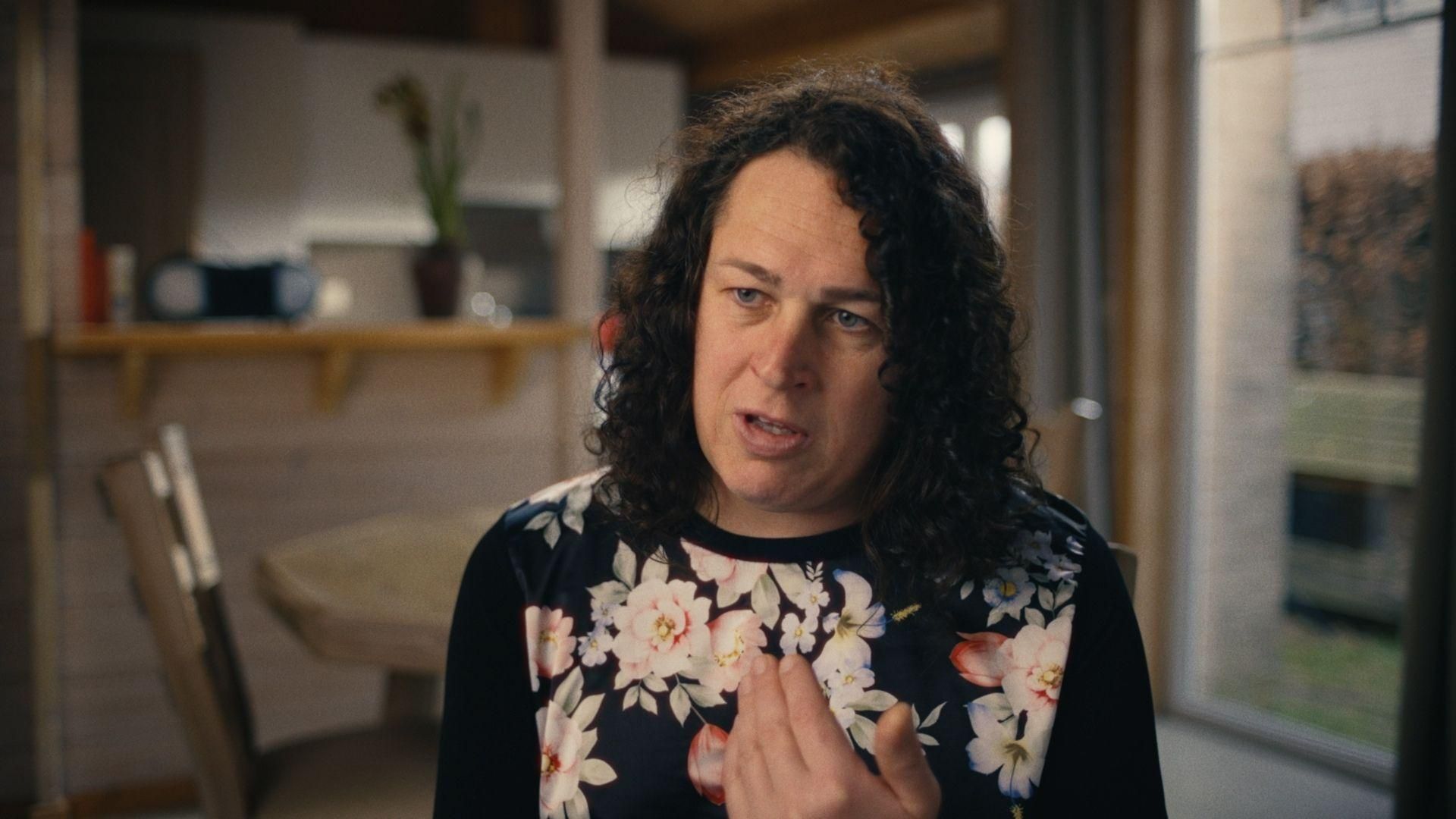 Jill Wuyts from Sanofi Belgium speaking expressively in a home setting, wearing a black floral top with a blurred living area in the background.