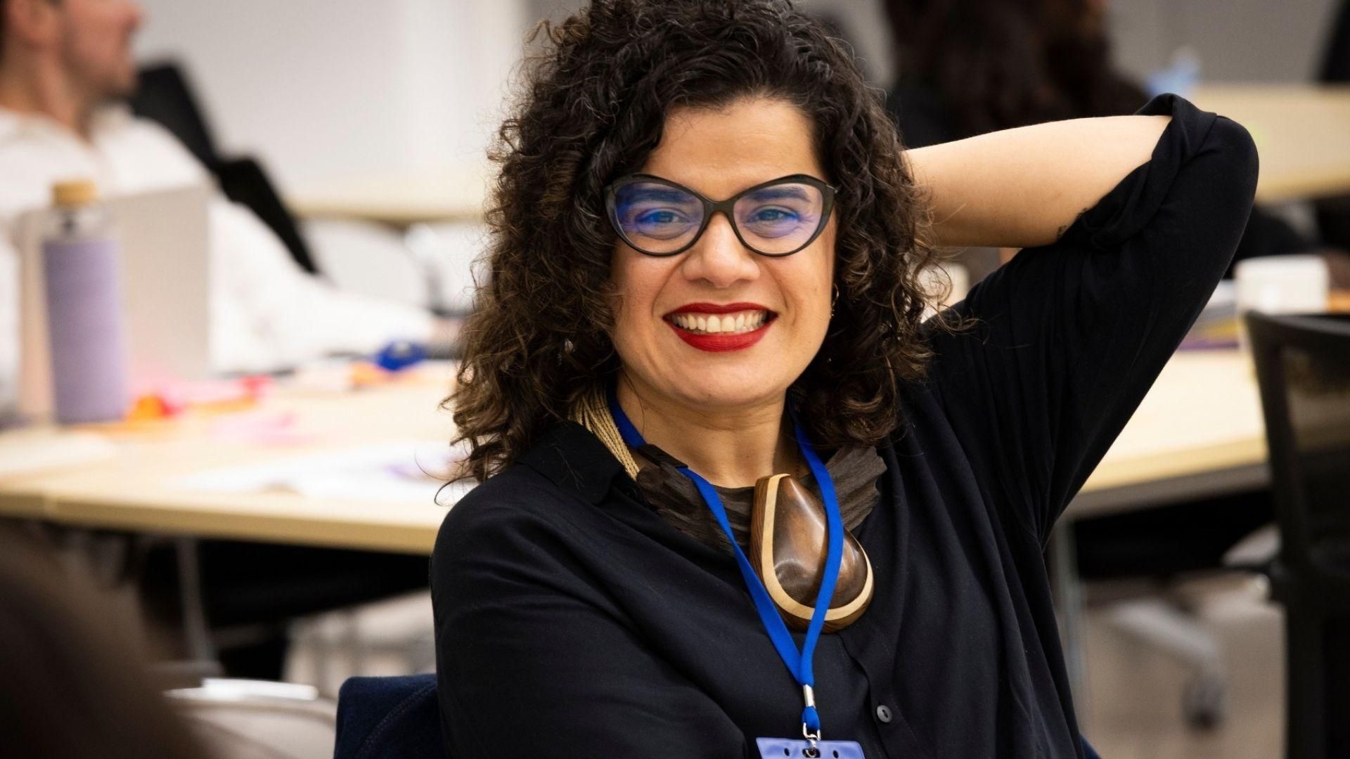 Neila Lopes, a woman with curly hair, sitting at a workshop table and smiling warmly. 