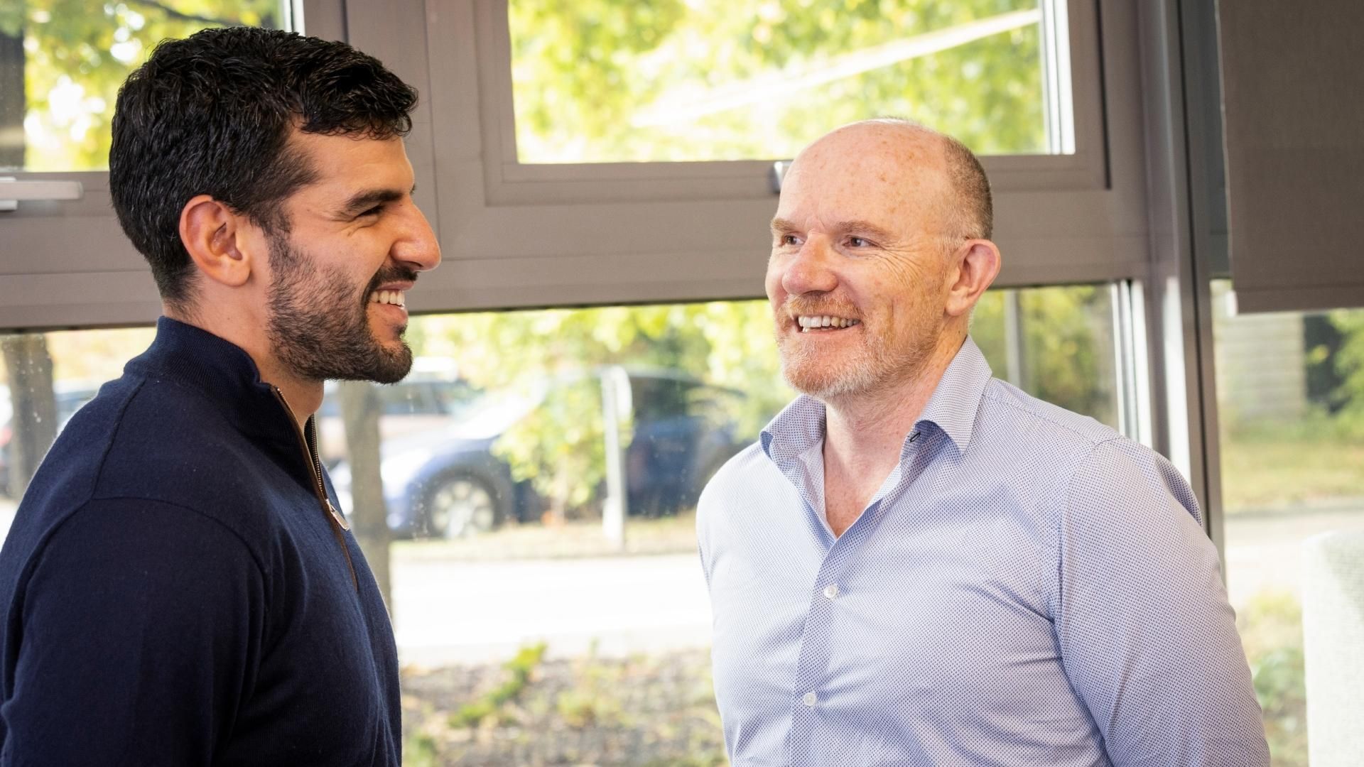 Two men in professional attire standing by a window, smiling and talking to each other. 
