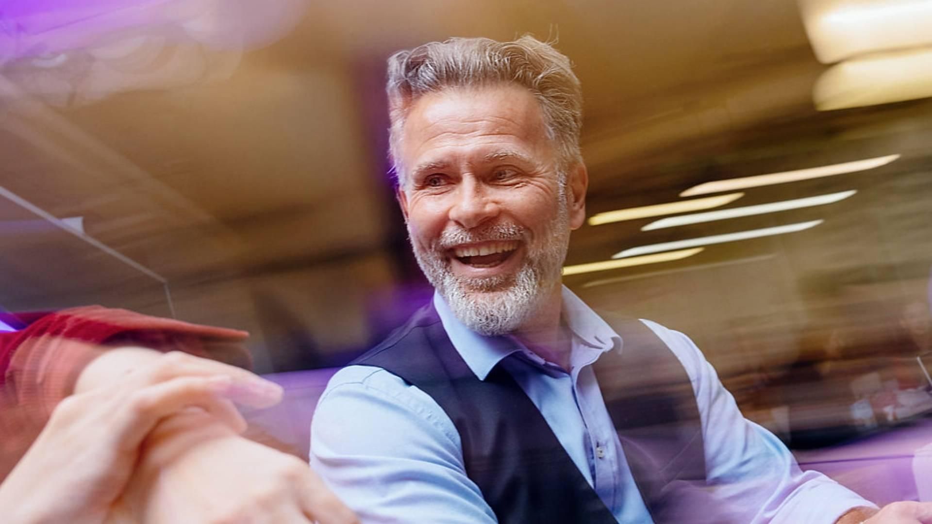 Person seated at a table indoors, wearing a light blue shirt and a dark vest. Another hand is visible in the foreground. The image features motion blur and soft purple and golden lighting effects.