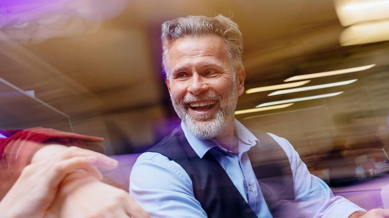 Person seated at a table indoors, wearing a light blue shirt and a dark vest. Another hand is visible in the foreground. The image features motion blur and soft purple and golden lighting effects.