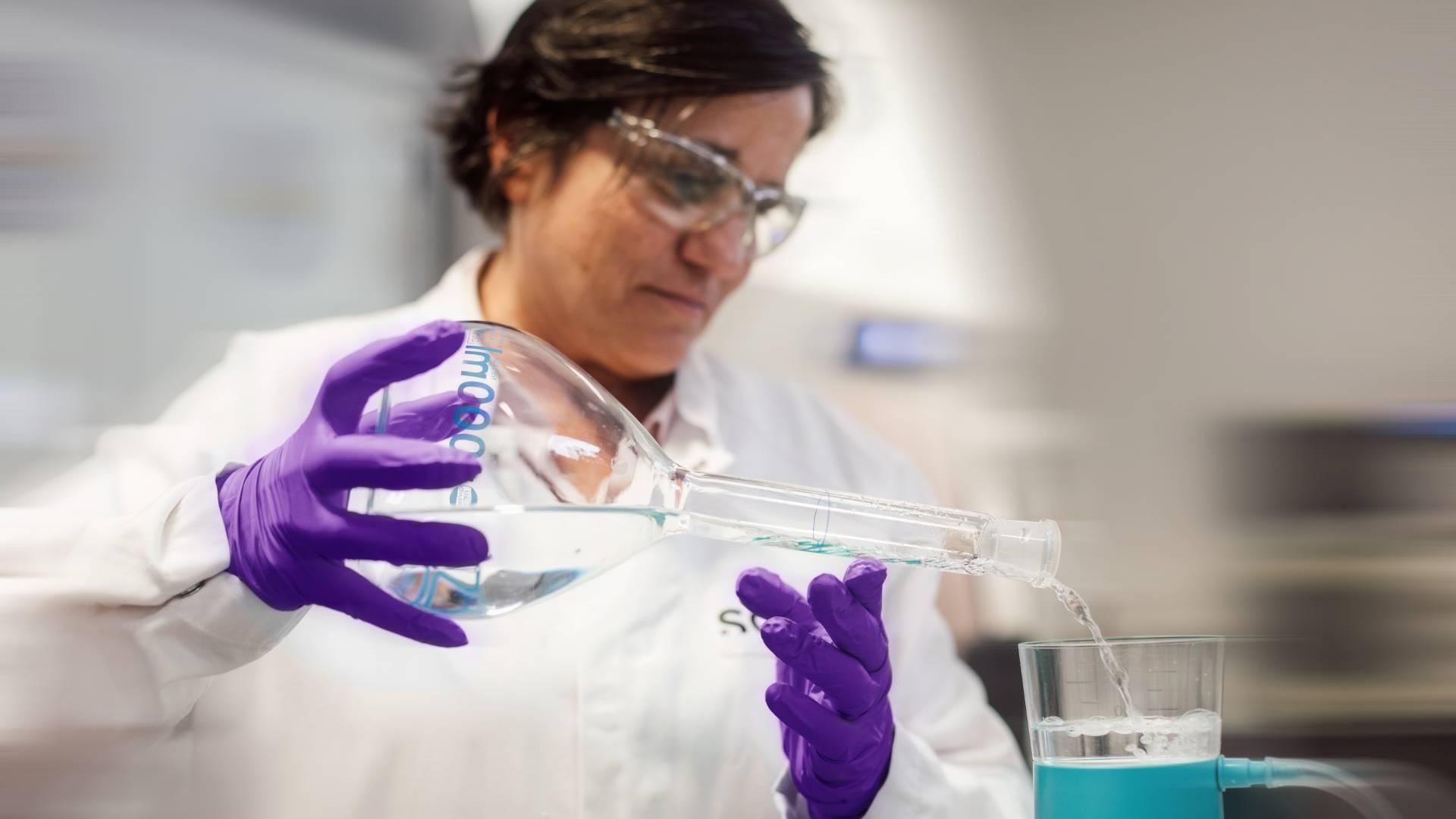Person wearing a lab coat, safety glasses and gloves, pouring liquid into a filtration device in a laboratory.