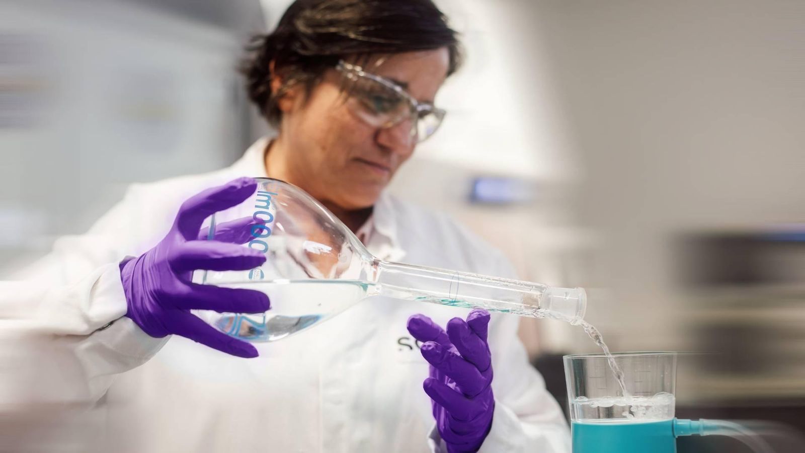 Person wearing a lab coat, safety glasses and gloves, pouring liquid into a filtration device in a laboratory.