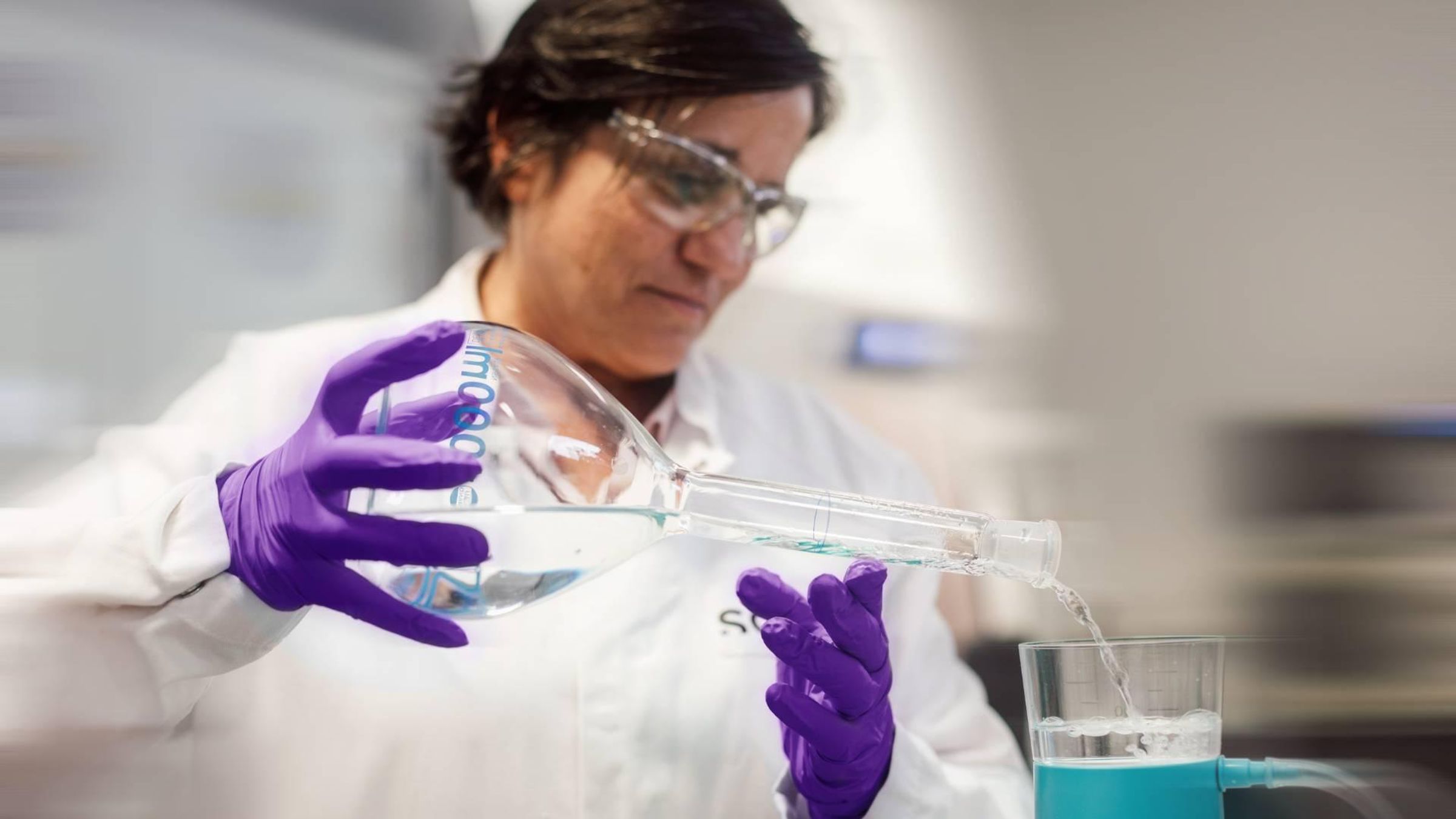 Person wearing a lab coat, safety glasses and gloves, pouring liquid into a filtration device in a laboratory.