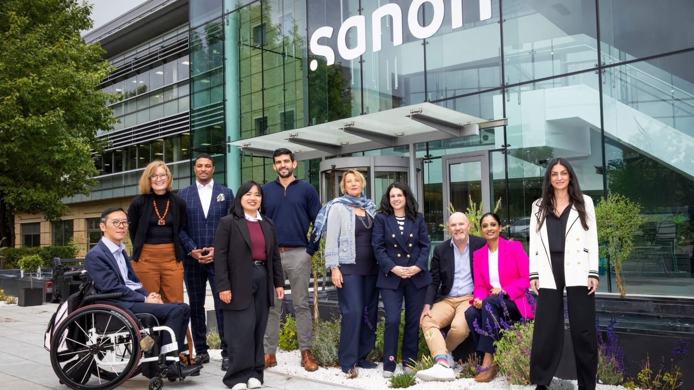 A diverse group of ten professionals in business-casual attire poses for a portrait in front of a modern glass building featuring the Sanofi logo.