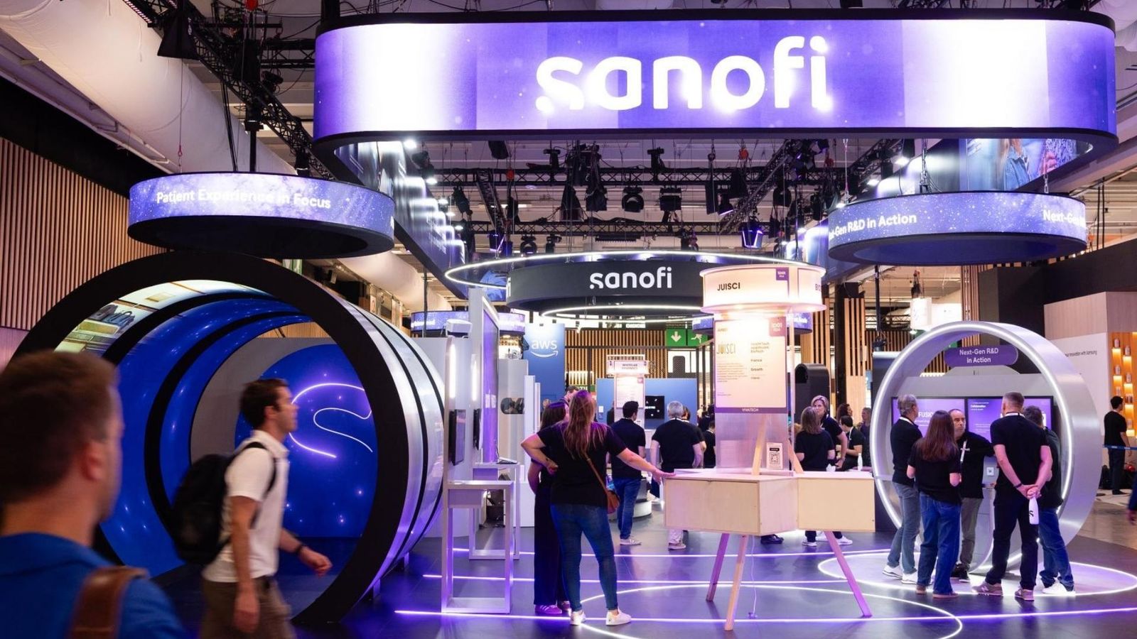Sanofi exhibition booth at VivaTech 2026. Illuminated signage featuring 'sanofi' in white letters on a purple background at the top of the booth. Below, curved LED screens and circular displays showcasing the brand and the messages 'Patient Experience in Focus' and 'Next-Gen R&D in Action'. The booth floor features circular patterns illuminated with purple lighting. Several visitors and staff members interact with demonstration tables and digital displays.