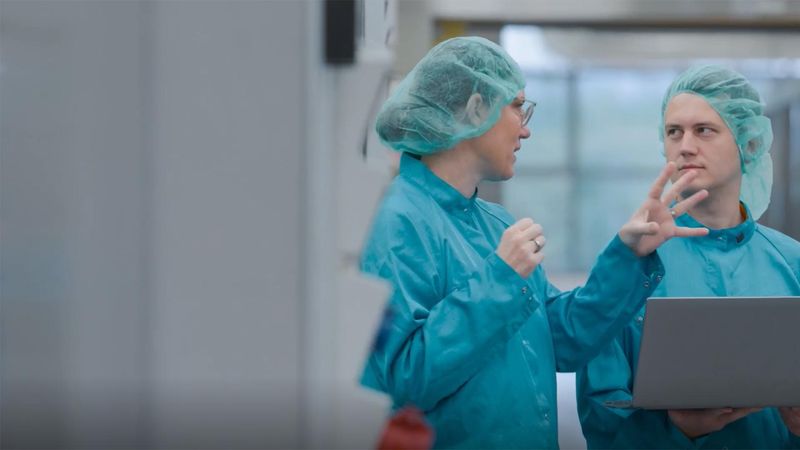 Two Sanofi scientists in lab coats and hairnets collaborating in a pharmaceutical manufacturing facility, one holding a laptop while discussing biopharmaceutical production processes, illustrating operational excellence and quality assurance across Sanofi's global supply chain.