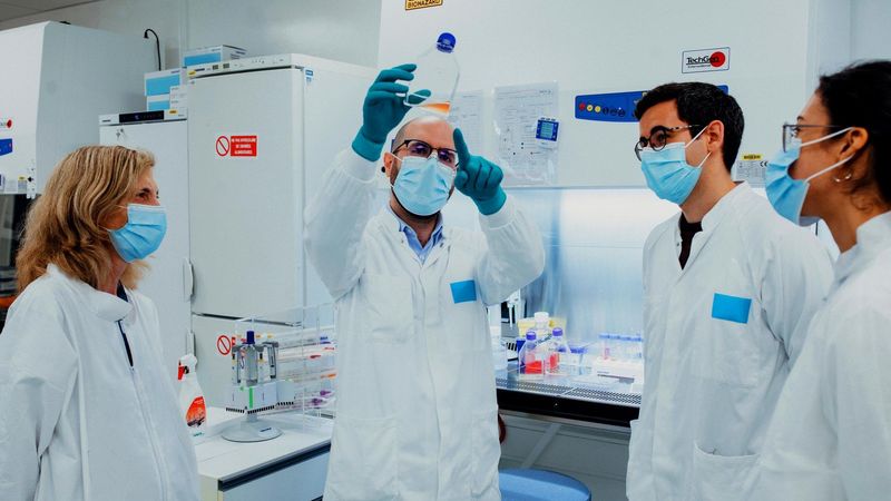 Two scientists, a lab assistant and a research clinician are in white lab coats, blue masks, and gloves and are working in a laboratory. One person holds a clear container with orange liquid while the others observe. The lab background includes a refrigerator, shelves with bottles and containers, and a workstation with scientific instruments.