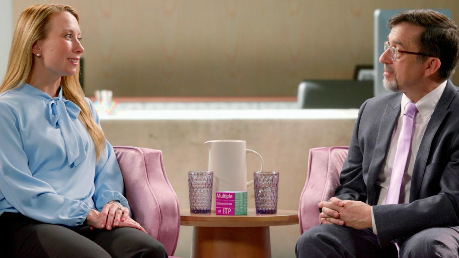 Holly, wearing a blue blouse, sits in an armchair in a living room setting with her provider, Dr. Michael Tarantino, wearing glasses, a grey suit, and a purple tie, during a conversation about ITP