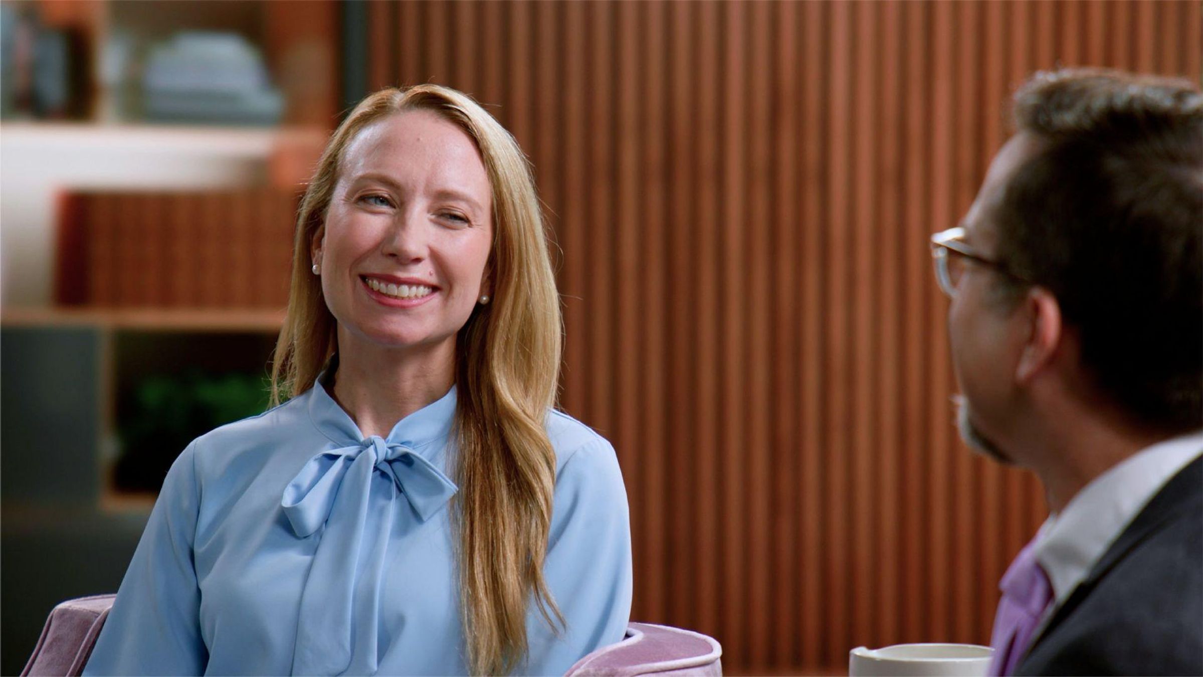 Holly, wearing a blue blouse, sits in an armchair in a living room setting, smiling at her provider, Dr. Michael Tarantino, who is wearing glasses, a grey suit, and a purple tie, to discuss her ITP management plan