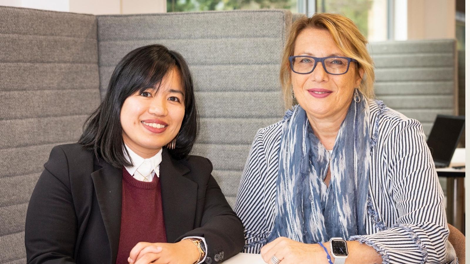 Two women sitting side by side in a modern office booth - both smiling warmly.