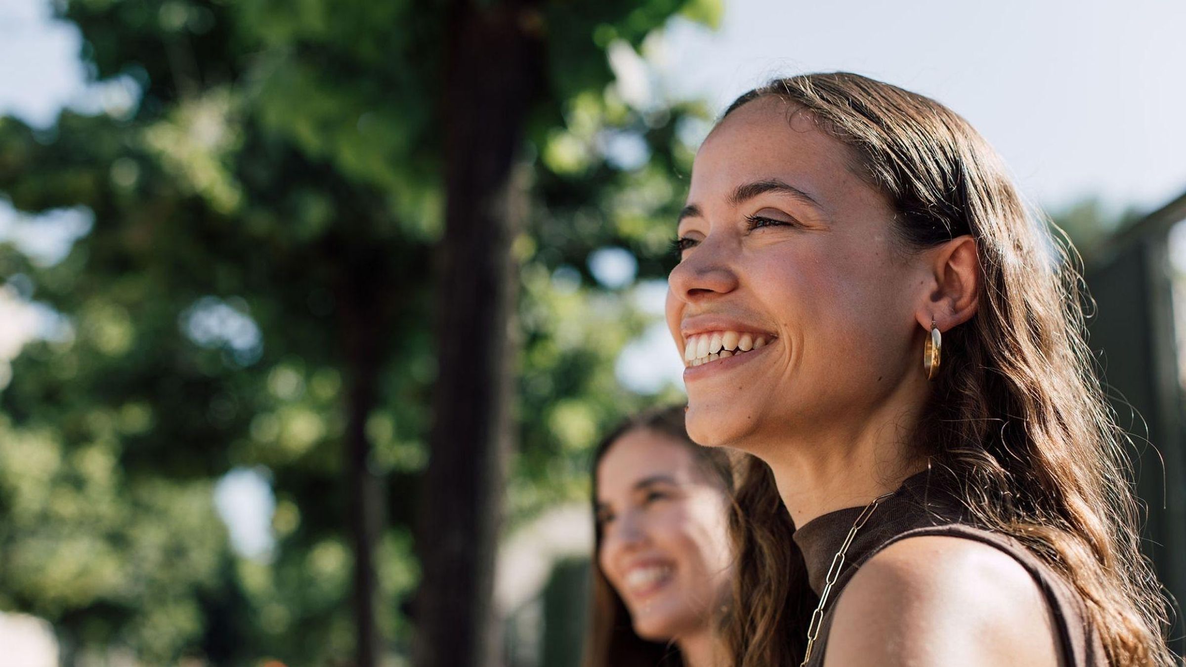 Two women laughing together outdoors in a sunny park with green trees in the background
