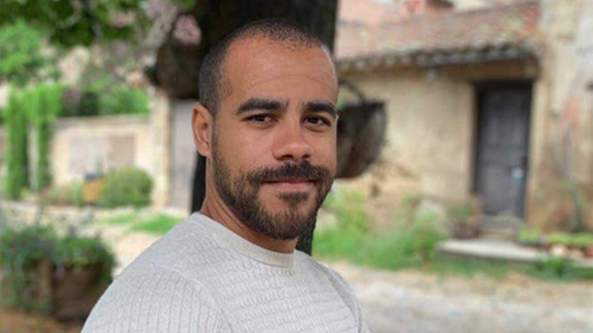 Bearded man in light-colored shirt standing outdoors in front of an old stone building with wooden door, surrounded by lush greenery