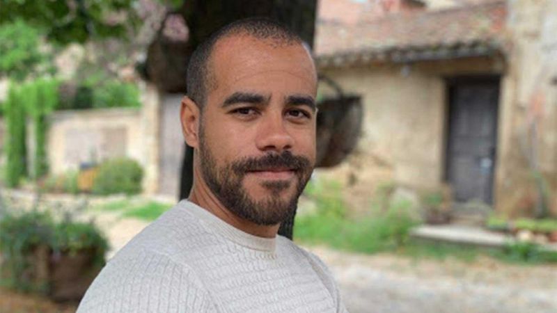 Bearded man in light-colored shirt standing outdoors in front of an old stone building with wooden door, surrounded by lush greenery