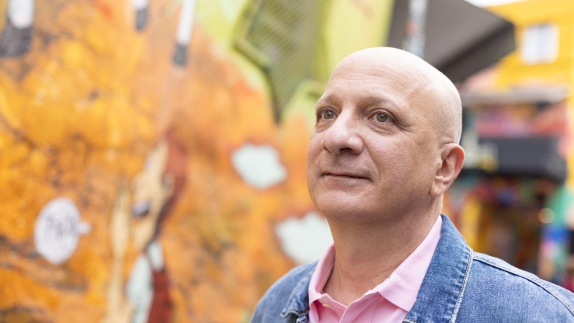 Pedro Pittella, with a shaved head, pink shirt and jean jacket, stands by a colorful wall in Sao Paulo