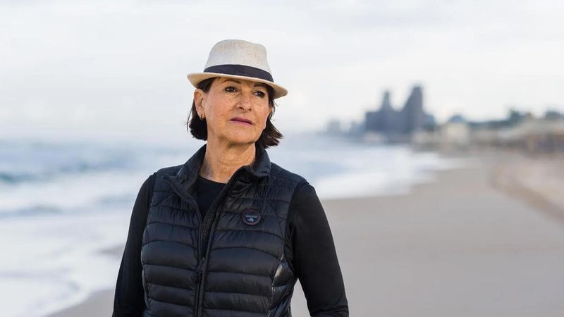 Woman standing on a beach, wearing a black puffer jacket and white hat, with ocean and urban coastline in the background