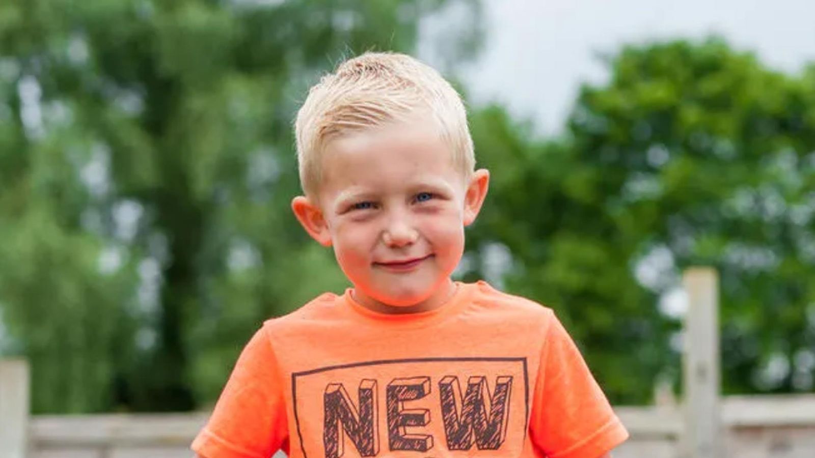 Alfie, a young blonde boy with acid sphingomyelinase deficiency, smiling and wearing an orange T-shirt with 'NEW' printed on it, in a green outdoor setting
