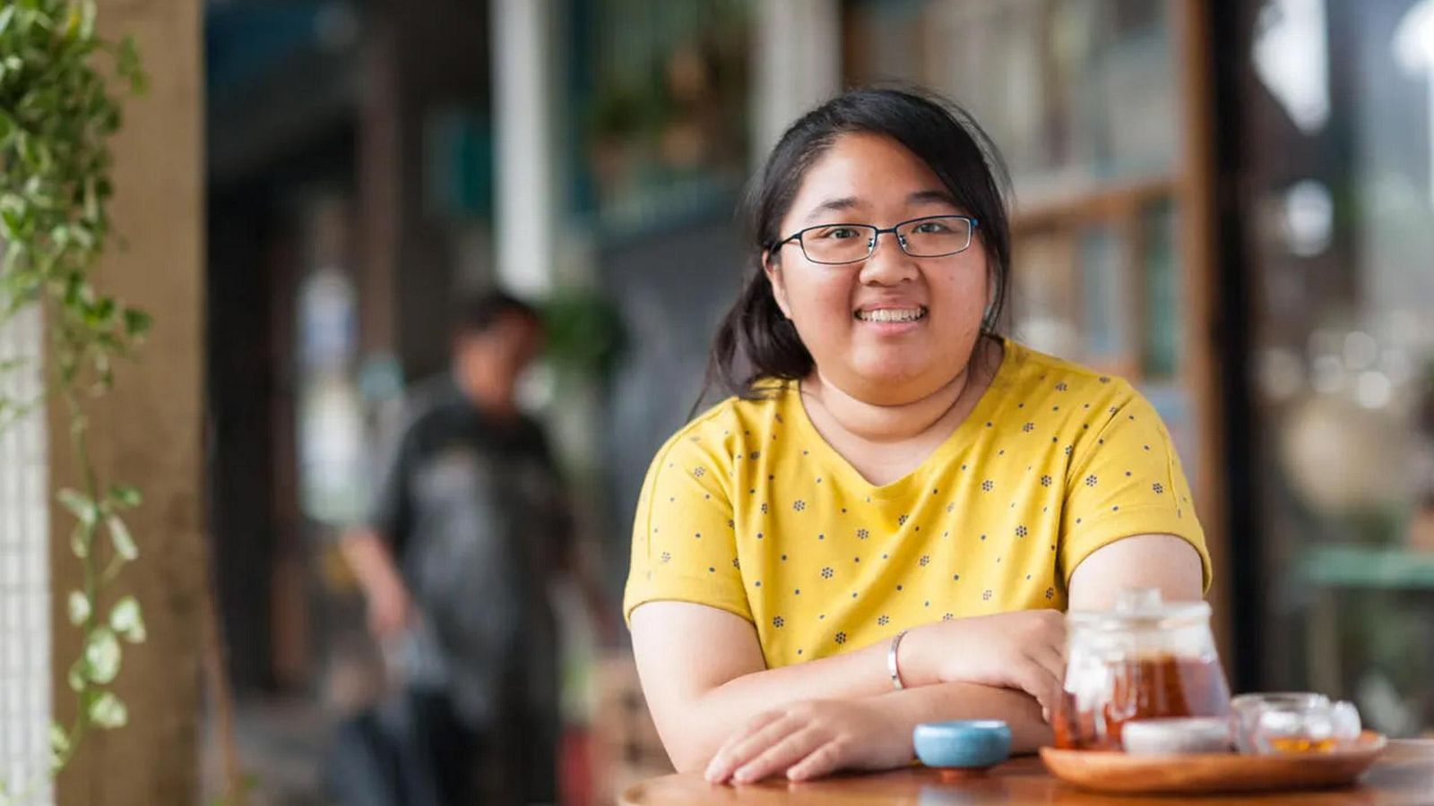 Hsieh, a Fabry disease patient from Taiwan, smiling, wearing glasses and a yellow patterned shirt, sitting at a table with a teapot and cup in front of her