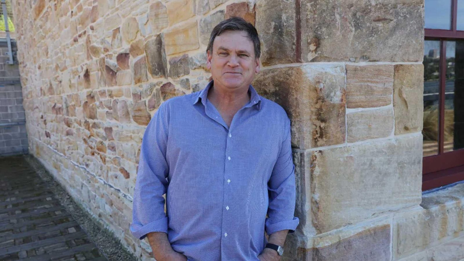 Ingo, a Pompe disease patient from Australia, standing against a stone wall, wearing a light blue shirt with his hands in his pockets