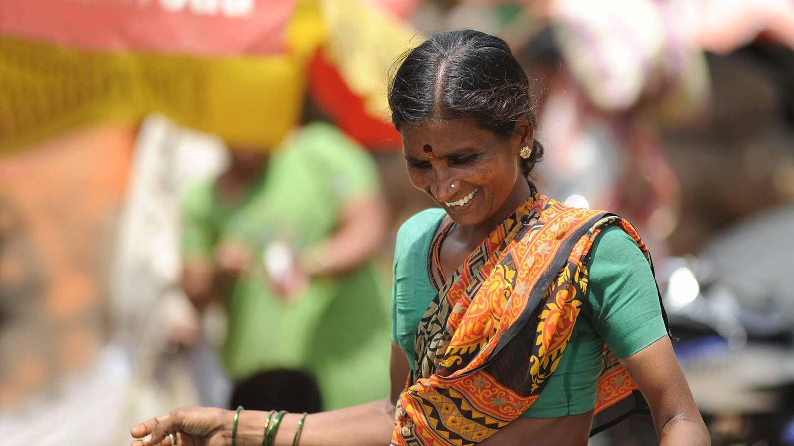 Portrait of a smiling Indian woman wearing a green blouse and orange-black dupatta, adorned with green bangles and a bindi, with other people in blurred background during an outdoor event