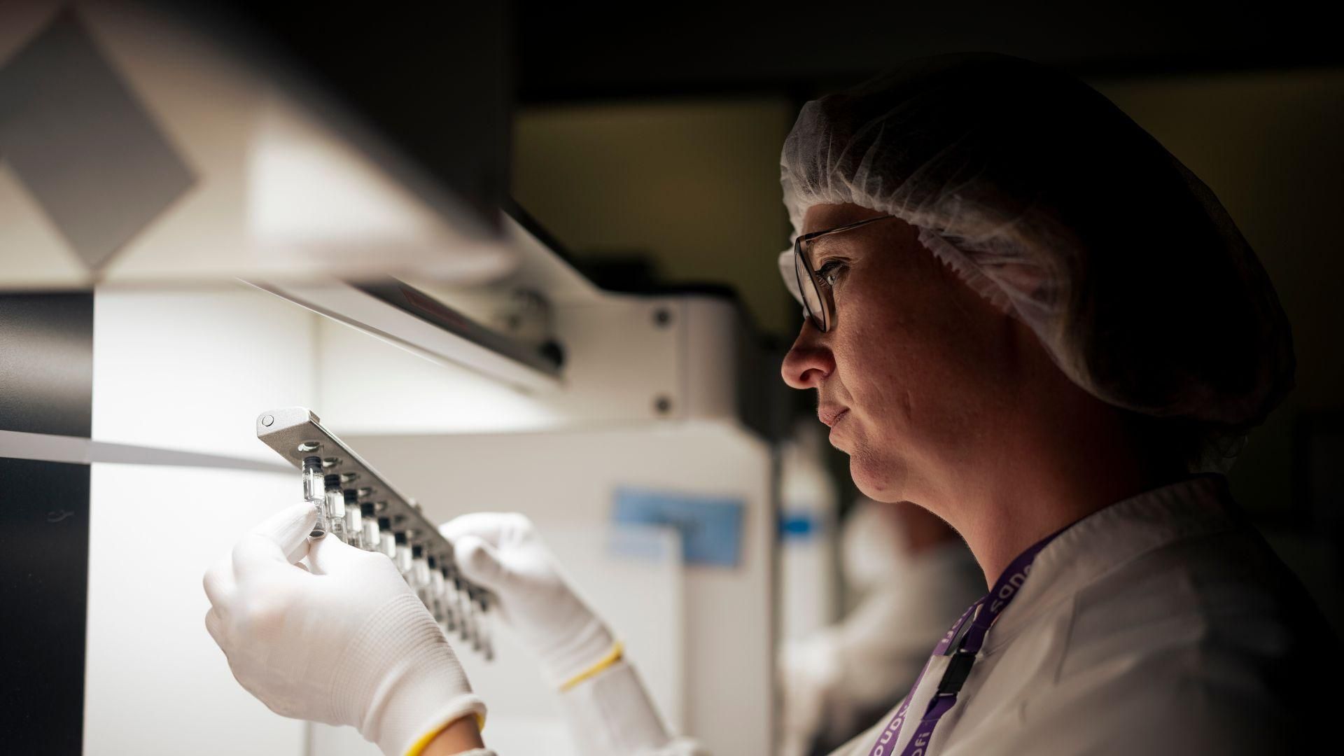 Nadège Bailleul inspects a set of 12 small clear vials on a steel rack next to a fume hood in a darkened room